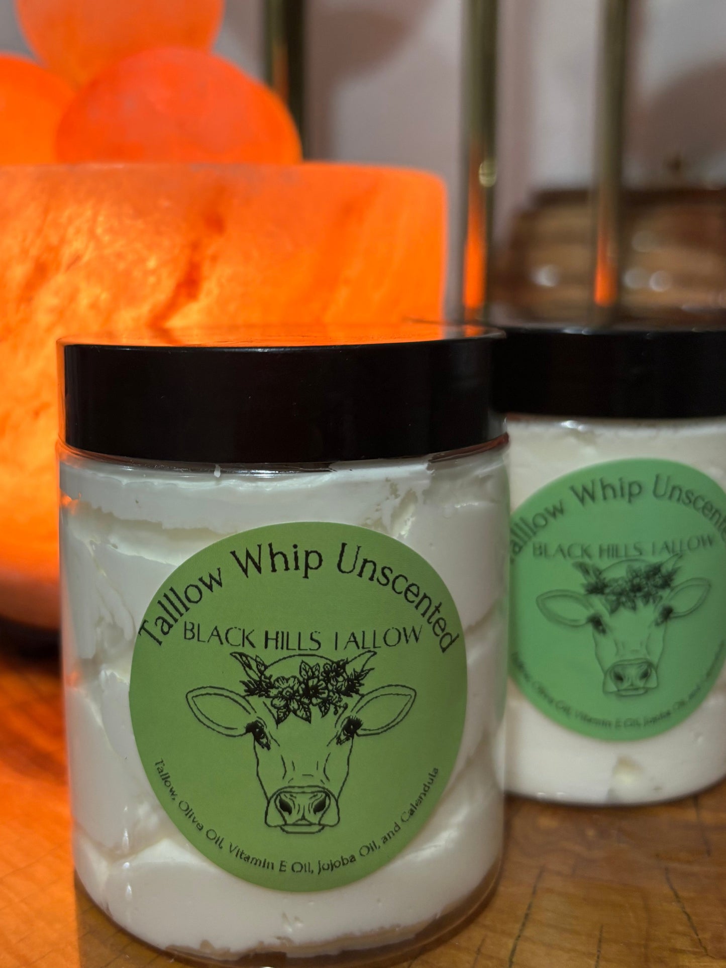 Two Black Hills Yallow candle jars in glass containers with black lids, featuring green labels with cow illustrations. The front jar is labeled 'Tallow Whip Unscented' and is in sharp focus, while a second matching jar sits behind it.