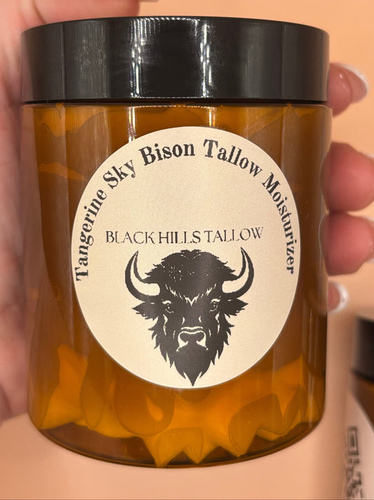A hand holding an amber glass jar candle with a black lid and a cream circular label featuring a black graphic of a bison and the text 'BLACK HILLS TAILWAY' and 'Tangerine Sky Tallow Moisturizer'.