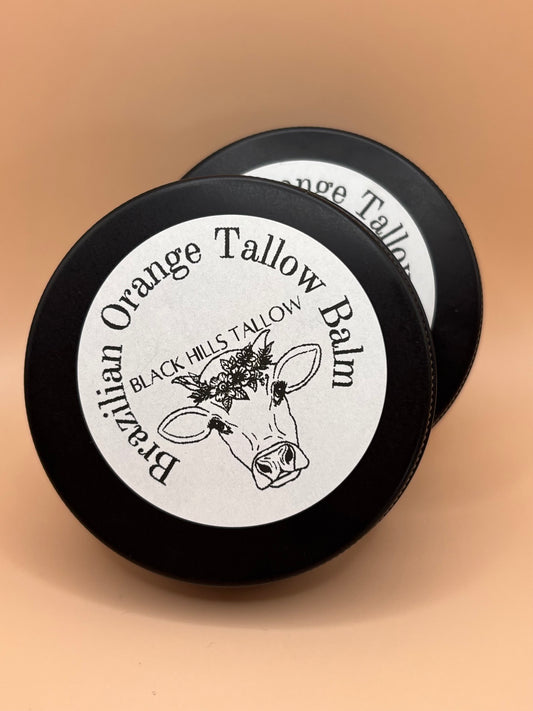 Two round black tins of Black Hills Tallow Brazilian Orange Tallow Balm, showing the white labels with a cow illustration, set against a solid peach background.